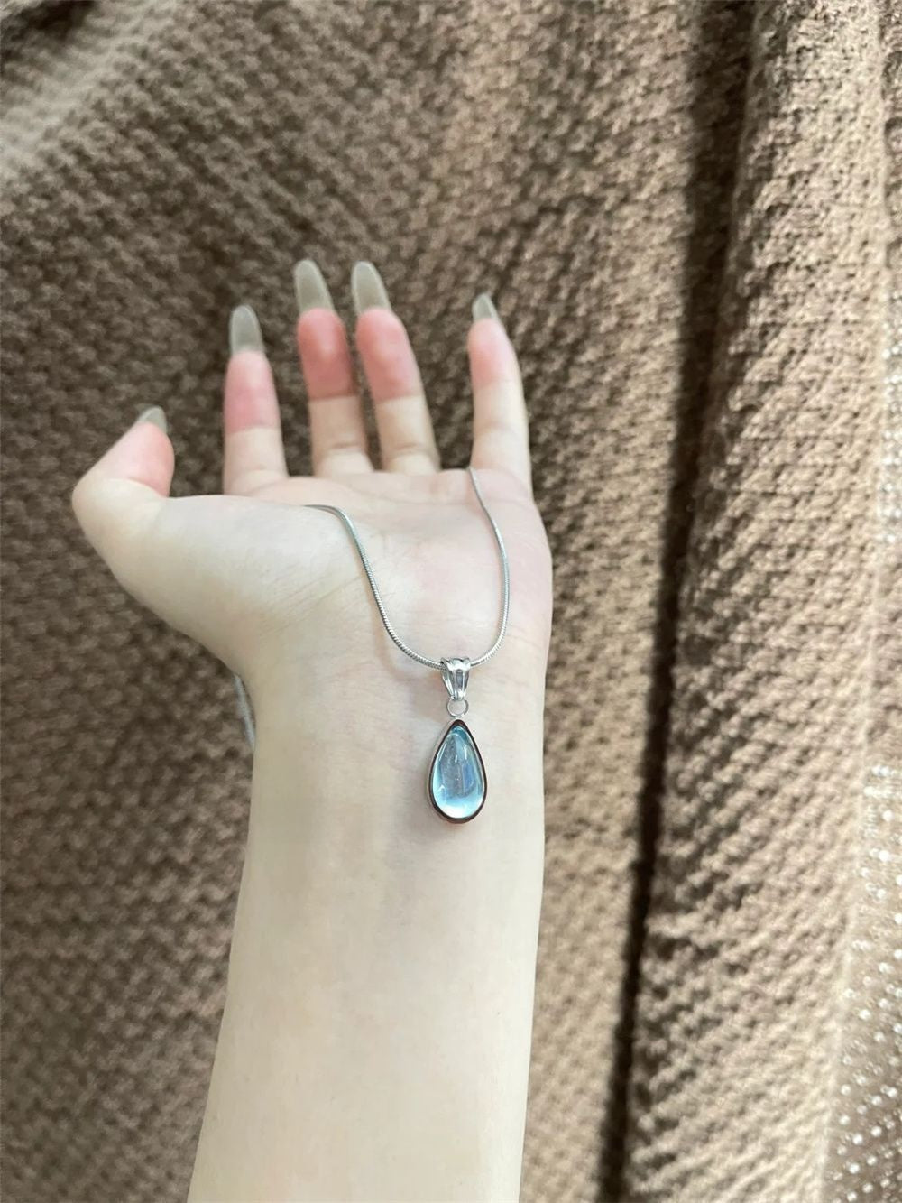 DewLume blue teardrop pendant and silver-tone chain displayed over a cream sleeve